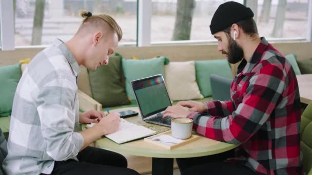 Před kamerou dva kancelářští pracovníci, kteří spolu pracují na projektu pomocí notebooku a dělají si poznámky do sešitu, zatímco sedí v kavárně — Stock video