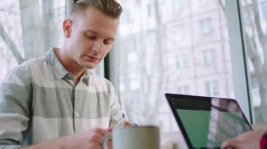 Closeup to the camera at the coffee shop with panoramic windows good looking man drinking some coffee and doing a college project make some notes. Shot on ARRI Alexa Mini