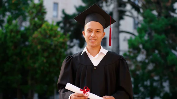 Graduado estudante feliz posando na frente da câmera com seu diploma no terno de graduação e boné — Fotografia de Stock