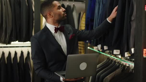 Afro American designer man working on his laptop he looking through the new collection of suits and make some notes on the laptop — Vídeo de Stock