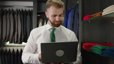 Modern kostüm dükkanı iyi görünümlü bir danışman. Dükkandaki giysi stoklarını kontrol etmek için dizüstü bilgisayar kullanıyor.