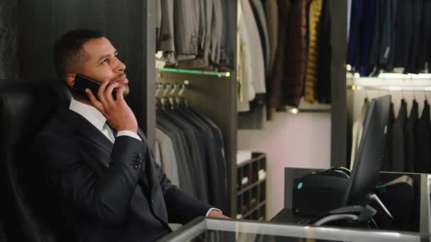 At the suit shop the consultant Afro American man have a conversation on his smartphone while sitting down on the office chair — Stockvideo