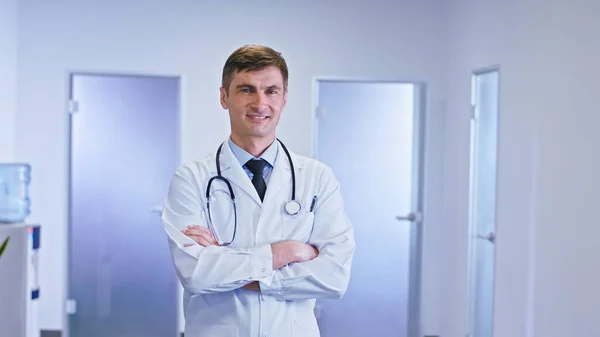 Перед камерой портрет врача в коридоре больницы, смотрящего прямо в камеру, у него скрещенные руки, у него стетоскоп на шее и большая улыбка. — стоковое фото