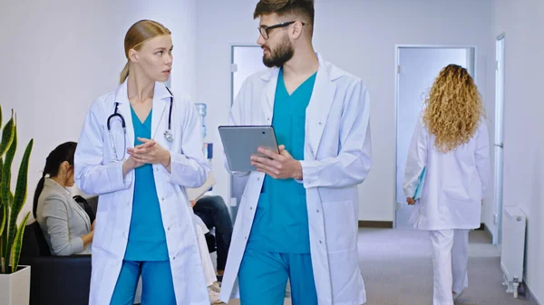 Moderní nemocniční chodba dva mladí lékaři multietnické ženy a muž kráčející před kamerou a diskutovat o budoucí léčbě pro pacienta při použití digitální tabletu koncept — Stock fotografie