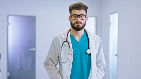 Skvěle vypadající chlap doktor v moderní nemocniční chodbě s brýlemi dívá přímo do kamery se soustředil nosí na krku stetoskop — Stock fotografie
