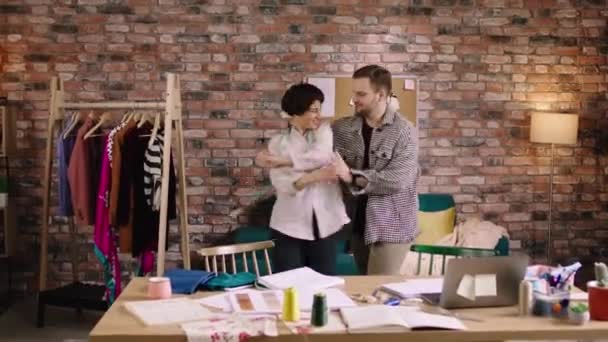 Two tailors are ecstatic to work together as they are dancing around to the music and having fun together in a office filled with sewing equipment — ストック動画