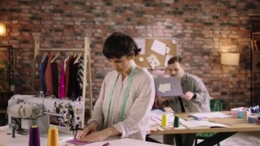 A woman with short hair is doing work at a sewing machine while a man behind her is working at a table answering phone calls and writing out designer notes. Shot on ARRI Alexa Mini
