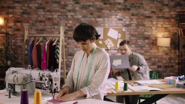 A woman with short hair is doing work at a sewing machine while a man behind her is working at a table answering phone calls and writing out designer notes. Shot on ARRI Alexa Mini — Video Stock