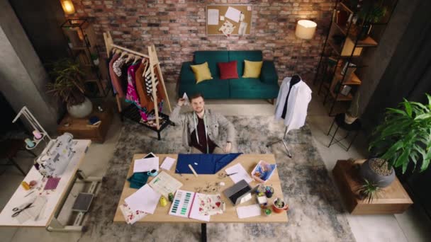 Top view. A man is in a very modern and hippie looking designer office, he is throwing a paper airplane in the air and doing some fabric work at his table, while enjoying the music playing — Vídeo de Stock