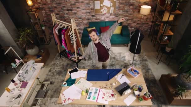 Στο πάνω μέρος. Χορεύοντας ενθουσιασμένος σχεδιαστής άνθρωπος στο σπίτι στο σαλόνι του εργάζεται πάνω από τη νέα του συλλογή από ρούχα που χάνουν τη μουσική από ακουστικά και κοιτάζοντας πάνω από τα σκίτσα — Αρχείο Βίντεο