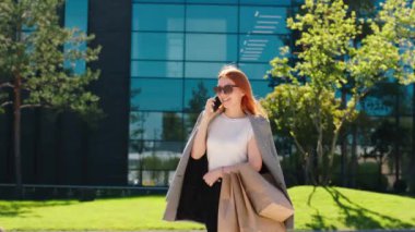 Güzel kızıl saçlı kadın, elinde alışveriş torbalarıyla kameranın önünde yürüyor ve güneş gözlüğü takan biriyle akıllı telefonuyla konuşuyor.