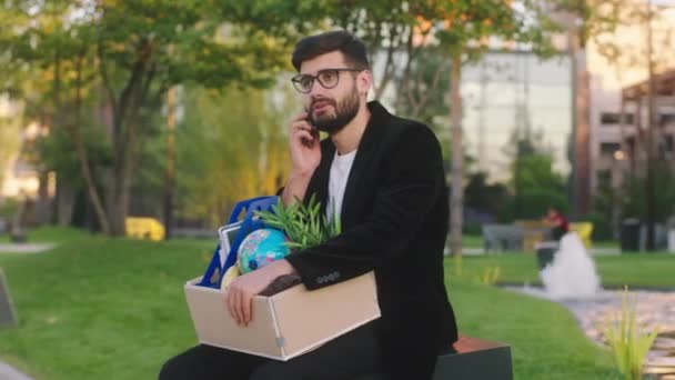 Handsome office manager with a office stuff in the box was fired from work he take a sit in the business centre park and speaks with someone on the phone. 4k — Vídeo de stock