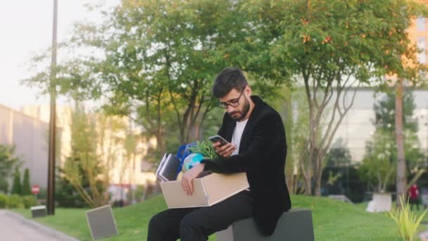 Good looking office manager young man very sad was fired from work he take a sit in the business centre park and typing something on the smartphone while holding his office stuff in the box — Αρχείο Βίντεο