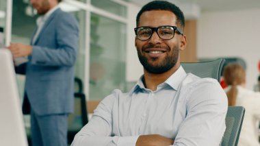 Çok çekici bir yönetici büyük ofisindeki masasında oturuyor siyah gözlüklerle dizüstü bilgisayarda çalışırken arkada yazıcının başında başka bir adam var.