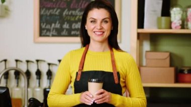 In front of the camera amazing barista woman with a perfect smile posing in front camera she holding a eco cup of coffee in the cozy coffee shop