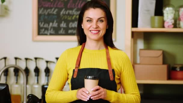 In front of the camera amazing barista woman with a perfect smile posing in front camera she holding a eco cup of coffee in the cozy coffee shop — Video Stock