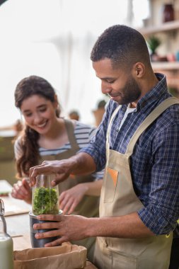 Karizmatik Afro-Amerikan adam ve kız arkadaşı Avrupalı mutfak adasına bakıyorlar birlikte sağlıklı kahvaltı hazırlıyorlar karıştırıcıya biraz taze meyve ekliyorlar ve sağlıklı bir karışım hazırlıyorlar.