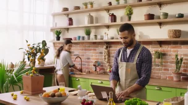 I det moderna köket man och hustru har en bra tid tillsammans kvinna diska medan hennes man arbetar på laptop på köksön och laga middag — Stockvideo