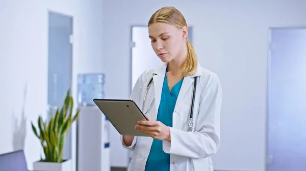 漂亮的、脸蛋集中的女医生用数码平板电脑记录病人的一些情况，然后直接看着医院走廊里的摄像机 — 图库照片