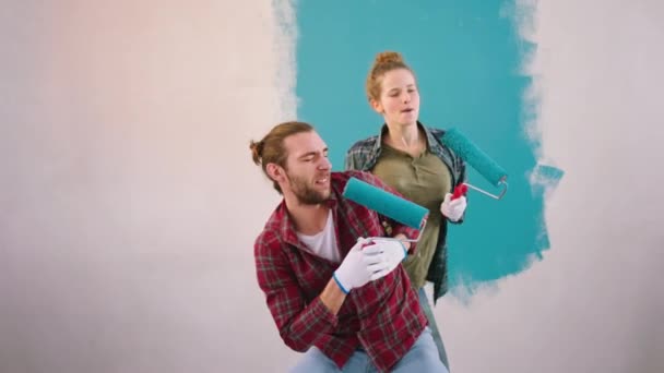 Joven pareja guapa mientras pinta la pared en la sala de estar empezar a cantar con el rodillo cepillo de pared para cantar y bailar — Vídeos de Stock