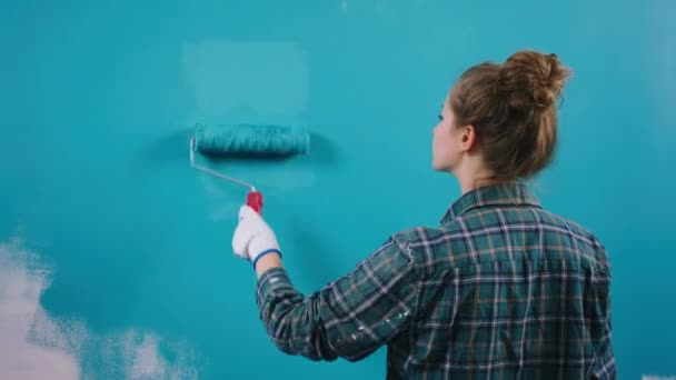 वर्किंग लेडी एक नीले रंग में पेंटिंग देख रही है एक बड़ी दीवार वह पेंट करने के लिए एक दीवार ब्रश रोलर का उपयोग कर — स्टॉक वीडियो