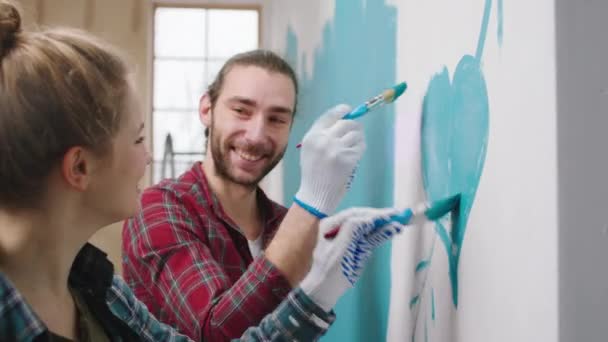 Souriant grand couple charismatique profiter du moment à la maison tout en peignant les murs ils font un grand cœur dans une couleur bleue avec de petites brosses et étreindre l'autre avec amour — Video