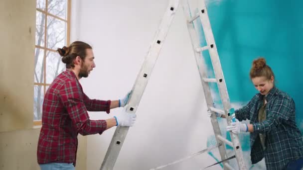 Pareja caucásica feliz y emocionada moviendo la escala para empezar a pintar las paredes en un color azul que sonríen grandes y se sienten emocionados — Vídeos de Stock