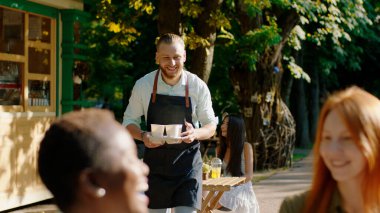 Parkın ortasında, kahve terasında, etnik çeşitliliğe sahip bayanlardan oluşan bir grup, birlikte vakit geçirmenin keyfini çıkarıyorlar, barista taze kahve getiriyor, kadınlar kahveyi alıyor ve konuşmaya başlıyorlar, gülümsüyorlar ve hissediyorlar.