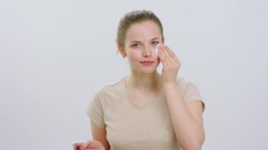Güzel bir kadın kameranın önünde yüzünü temizlemek için makyaj diski kullanıyor tatlı tatlı gülümsüyor ve beyaz arka plandaki kameraya bakıyor. 4k