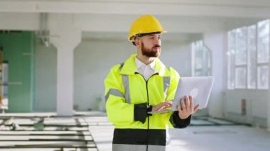 İnşaat alanında kameraya doğru yürürken aynı zamanda dizüstü bilgisayarda çalışırken aynı zamanda inşaat planını analiz etmek için dizüstü bilgisayarı tutuyor. ARRI Alexa Mini 'de çekilen.