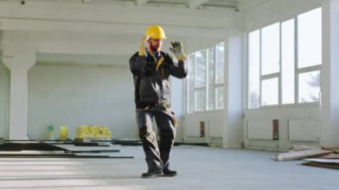 Güvenlik kaskı ve üniformalı çekici genç mühendis modern inşaat binasında iş günü sonunda heyecanlı ve mutlu bir şekilde dans ediyor. 4k