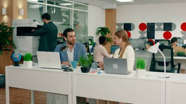 Yakışıklı bir adam ve bir kadın çalışma odasında kablosuz kulaklık takmış, koltuklarında oturuyorlar, arkada geniş omuzlu bir adam fotokopi makinesinin başında.