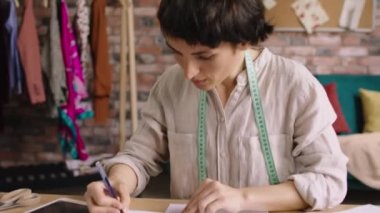 Modern terzilik atölyesi kadın giyim tasarımı defterine yeni bir taslak çizer. Sevgiyle dikmek istediği yeni elbisenin taslağını yapar. Zevk konsepti olan terzilik işini yapar.
