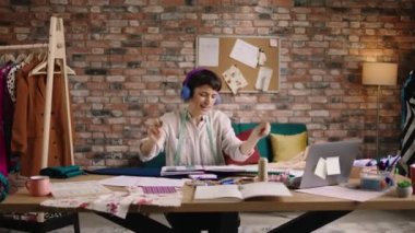 Gülümseyen güzel bayan moda tasarımcısı yeni koleksiyonundan bazı düğmeler alıyor ve moda ofisindeki iş yerindeki kulaklıklardan müzik dinlerken dans ediyor ve yavaşça hareket ediyor. 4k
