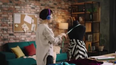 Kulaklıklardan müzik dinlerken dans eden karizmatik ve çekici terzi kadın ve terzideki müşterisi için gelecekte giyeceği elbise için bazı ölçümler yapıyor. Vuruldu.