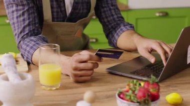 Mutfaktaki kameraya yakın, güzel gülüşlü, kredi kartını kullanarak bilgisayardan bir şeyler ödeyen mutlu adam. 4k