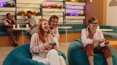 Üç güzel arkadaş mavi fasulye koltuklarında oturmuş video oyunu oynuyorlar. Arkalarından da üç adam onları izliyorlar..