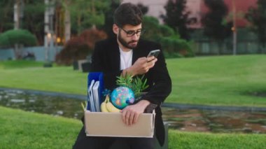 Ofis müdürünün önünde ofis eşyalarını kutuda tutarken ve akıllı telefondan bir şeyler yazarken iş merkezinin parkında otururken çok üzgün bir şekilde işten atıldı. 4k