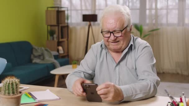 自宅の作業台に座っている老人のビデオを自宅のリビングルームで撮影し スマートフォンと笑顔を通して見ています — ストック動画