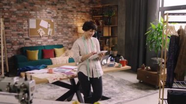 Elinde dijital tablet bulunan harika görünümlü bayan terzi, terzide giyecek koleksiyonu için bir taslak hazırlıyor.