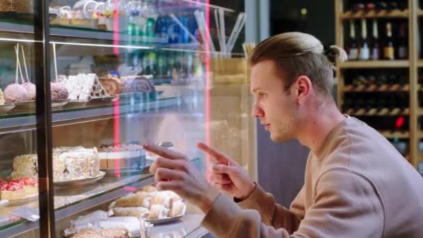 カメラに近いハンサムな若い男性の顧客は コーヒーショップのショーケース冷蔵庫から好きなデザートを選択します — ストック動画