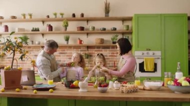 Büyük ve büyük bir modern mutfakta, büyük gülümsemeli iki küçük kız aileleriyle birlikte yemek pişirip birlikte yeşil salata yiyorlar. 4k