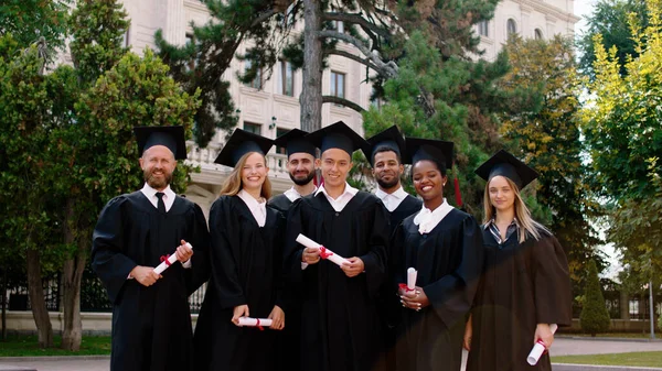 In the college garden graduation day group of charismatic and excited students posing in group together in front of the camera they wearing graduation suits and caps and smiling large. Portrait