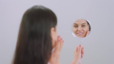 In front camera on white background woman holding a mirror and touching her perfect face looking through the mirror to the camera and smiling large. Shot on ARRI Alexa Mini
