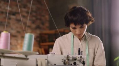 Modern çatı katı tasarımı atölyesi. Kısa saç terzisi olan harika bir kadın. Dikiş makinesine kıyafet dikiyor. ARRI Alexa Mini 'de çekilen