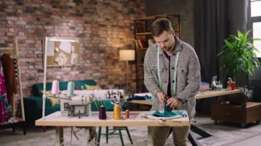 Modern daire stüdyosu yakışıklı adam moda tasarımcısı kumaşları kullanarak çalışma masasında yeni bir kıyafet koleksiyonu yapıyor. ARRI Alexa Mini 'de çekilen