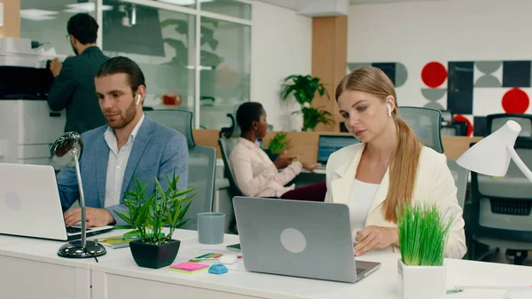 A beautiful blonde woman with her hair tied in a low ponytail, wearing a white blazer is doing work on her laptop and beside her is a handsome man in a blue suit also doing work on his laptop while