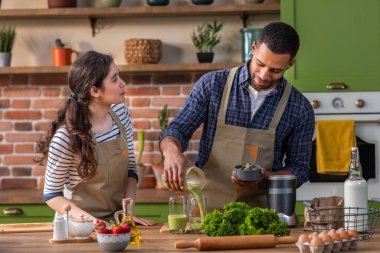Karizmatik Afro-Amerikan adam ve kız arkadaşı Avrupalı mutfak adasına bakıyorlar birlikte sağlıklı kahvaltı hazırlıyorlar karıştırıcıya biraz taze meyve ekliyorlar ve sağlıklı bir karışım hazırlıyorlar.