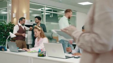 While a handsome smart man is talking on his wireless earphones at the office, a woman is beside him doing work while two intelligent looking men are having a conversation beside the printer and