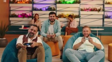 Biri diğerinden daha yaşlı iki adam birbirlerine karşı yarışıyor ve video oyunları oynuyorlar, mavi fasulye koltuklarında otururken diğeri onlara tavsiye veriyor.